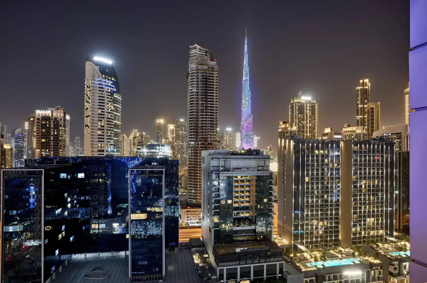 Night view of Dubai skyline featuring illuminated skyscrapers, including the Burj Khalifa lit with colorful lights.