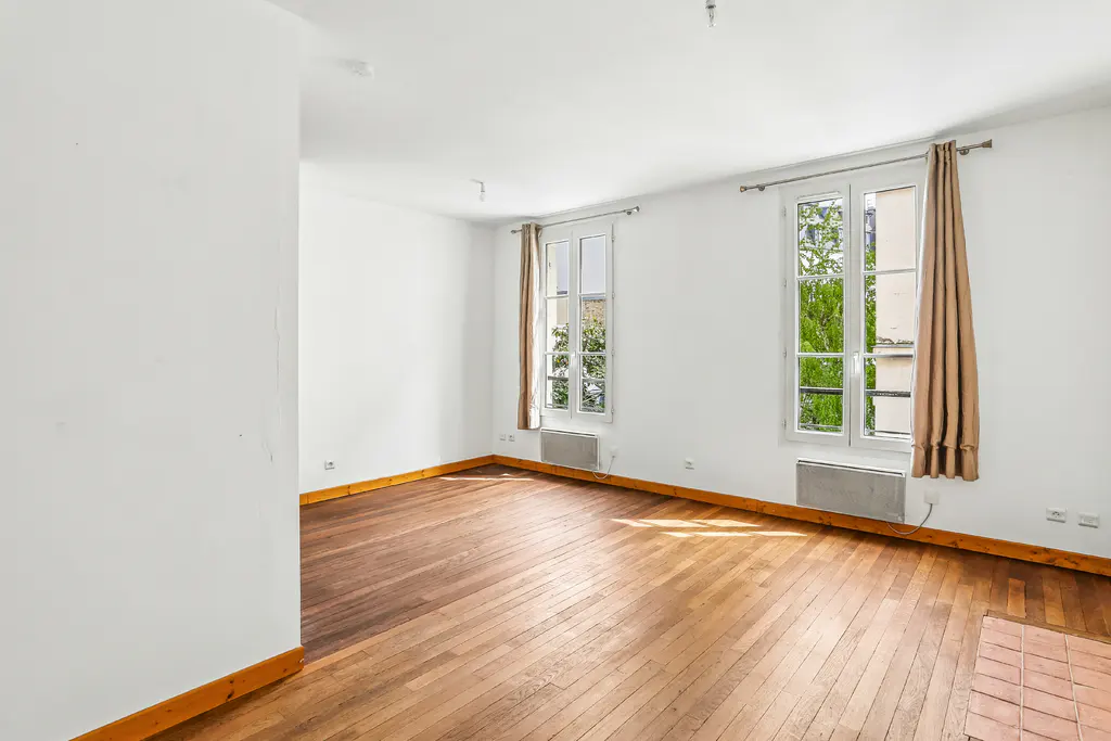 Bright, empty room with white walls, wood floors, and two windows with tan curtains.