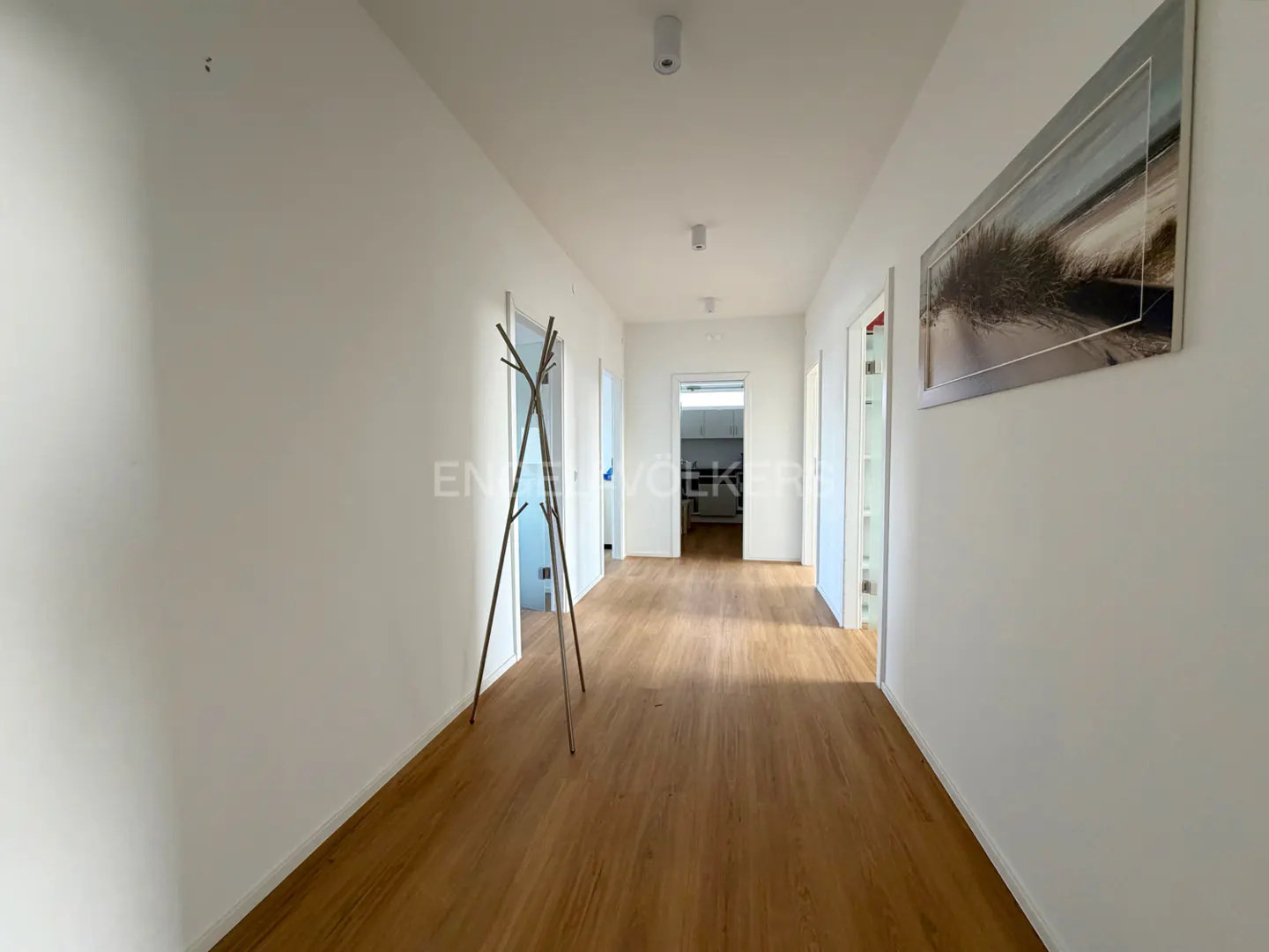 Hallway with wood floors, white walls, and a coat rack. A beach photo hangs on the right wall. A kitchen is visible at the end of the hall.
