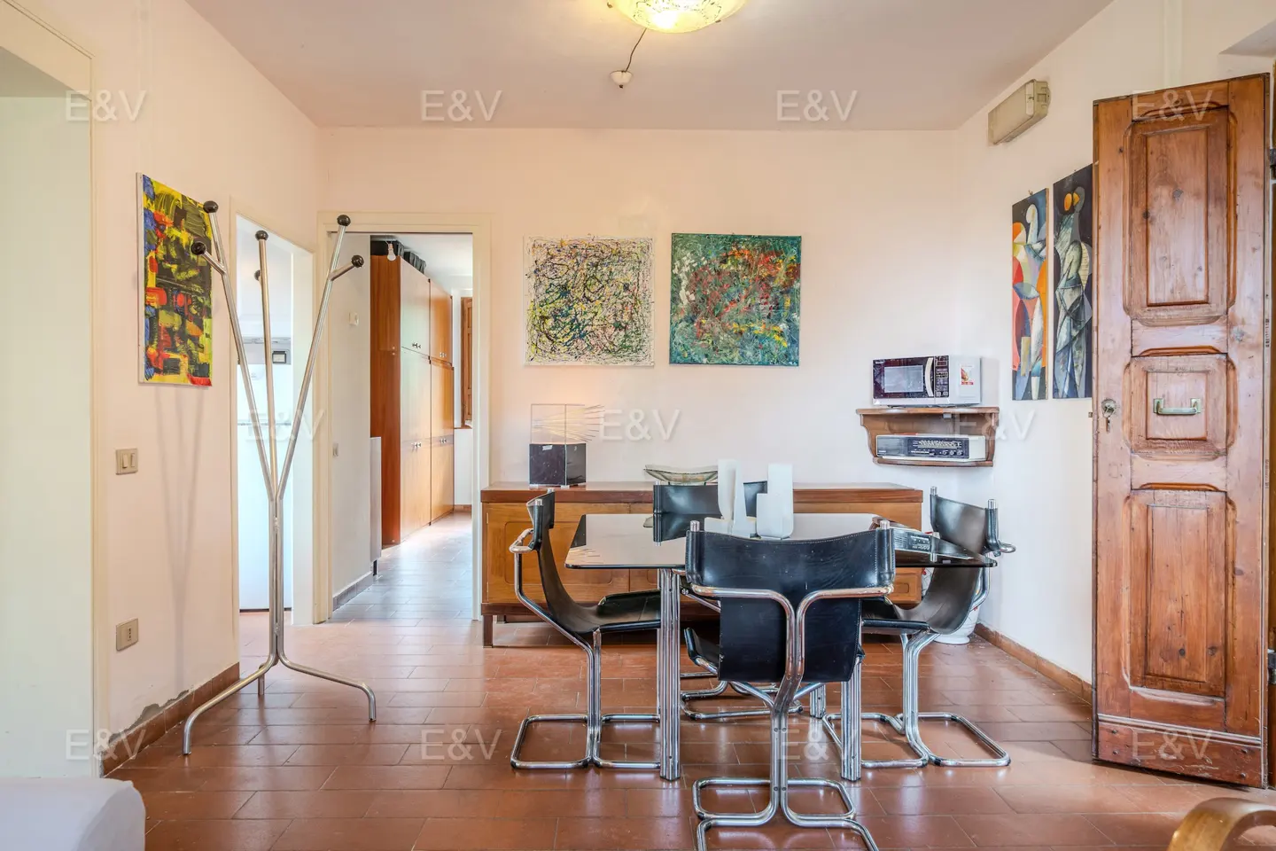 A dining area with a table, chairs, and artwork on the walls. The floor is tiled in a terracotta color.