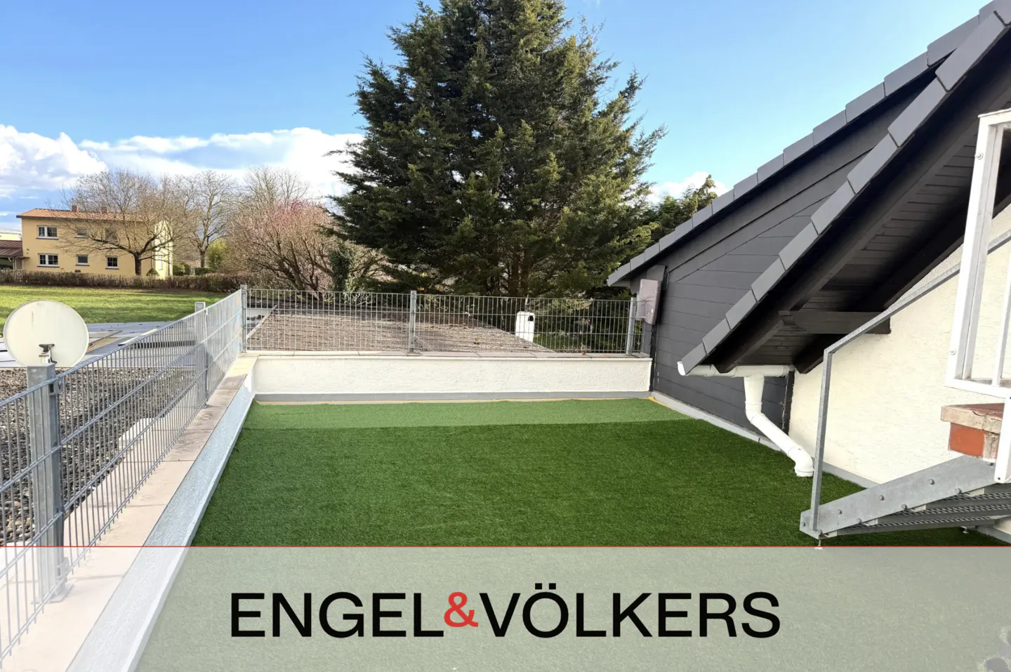 A rooftop terrace with green artificial grass, a metal fence, and a view of a building and trees under a blue sky.