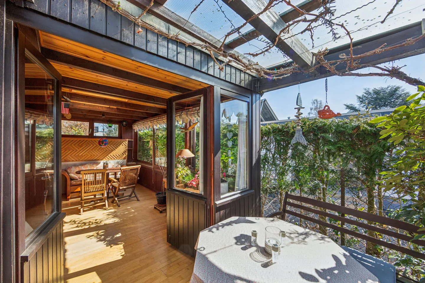 Sunroom with wood floors, table, and chairs. Outdoor patio with table, chairs, and greenery. Black trim and a clear roof.