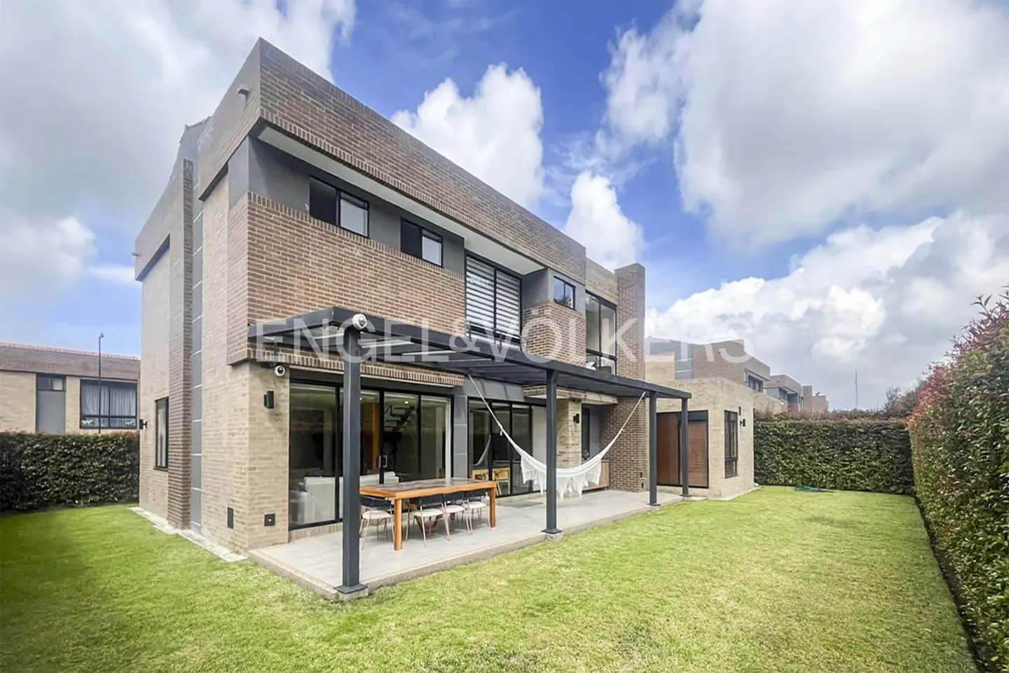Brick house with a patio, table, hammock, and green lawn under a cloudy sky.