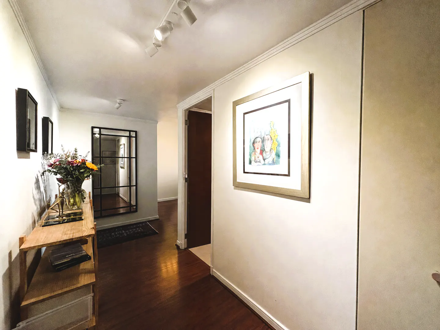 Hallway with hardwood floors, white walls, and track lighting. A console table with flowers sits near a mirror. Artwork hangs on the walls.
