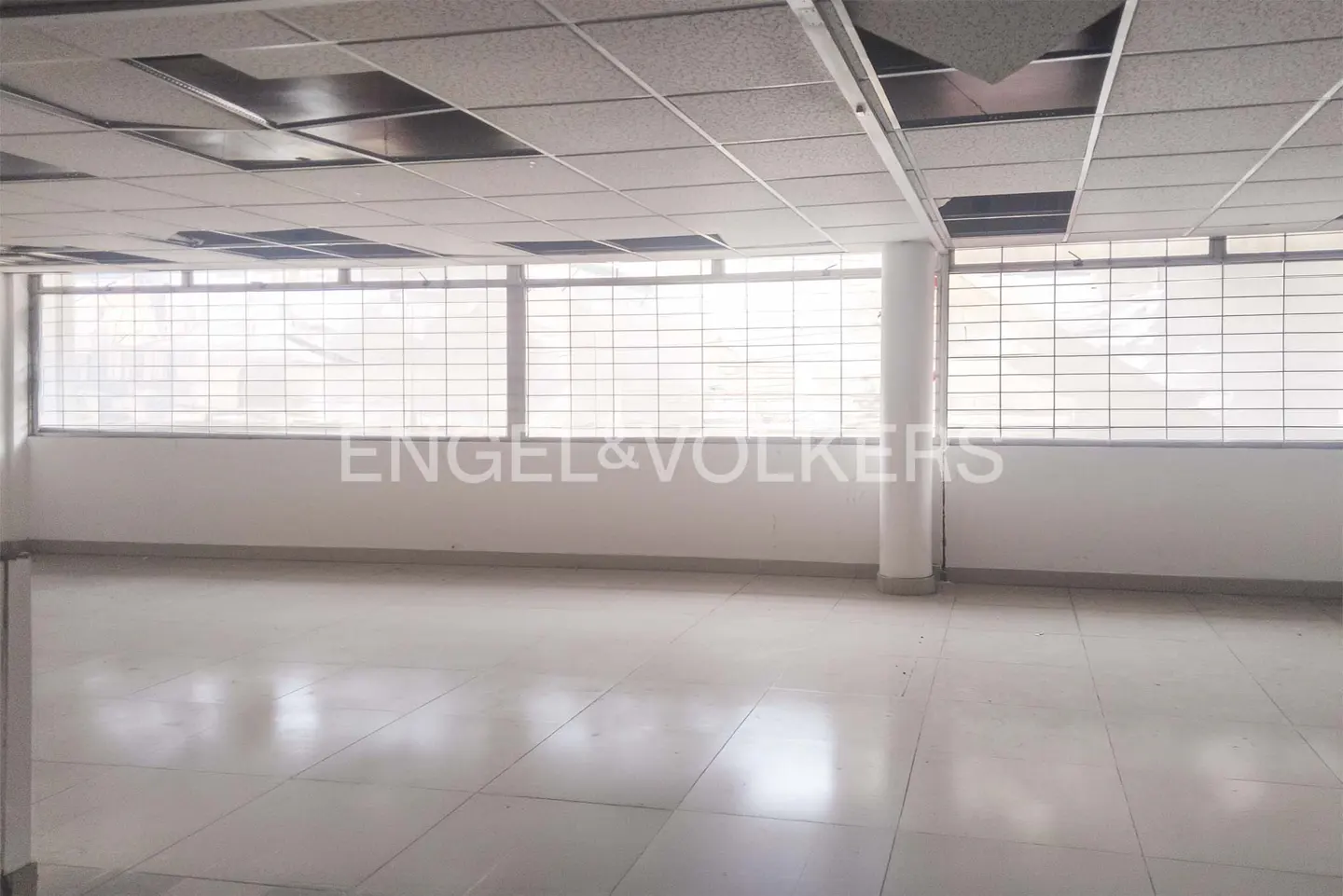Empty office space with white walls, tile floor, and large windows with metal bars. Ceiling tiles are visible, some missing. Engel & Volkers logo on wall.