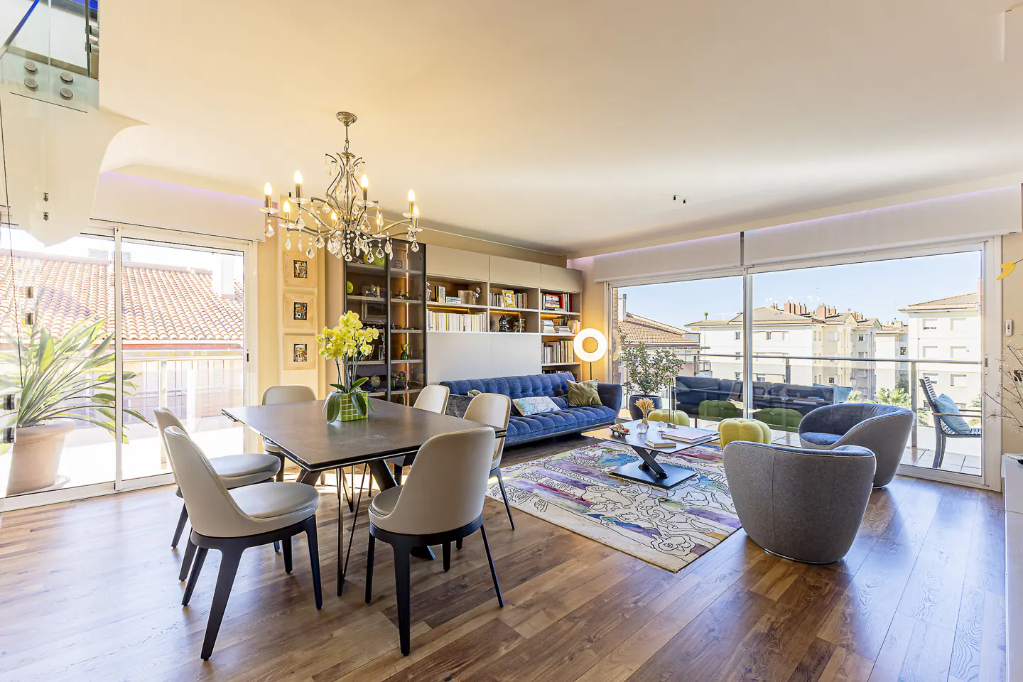 Bright, open-concept living and dining area with wood floors, large windows, and a blue velvet sofa. A chandelier hangs above a dining table.