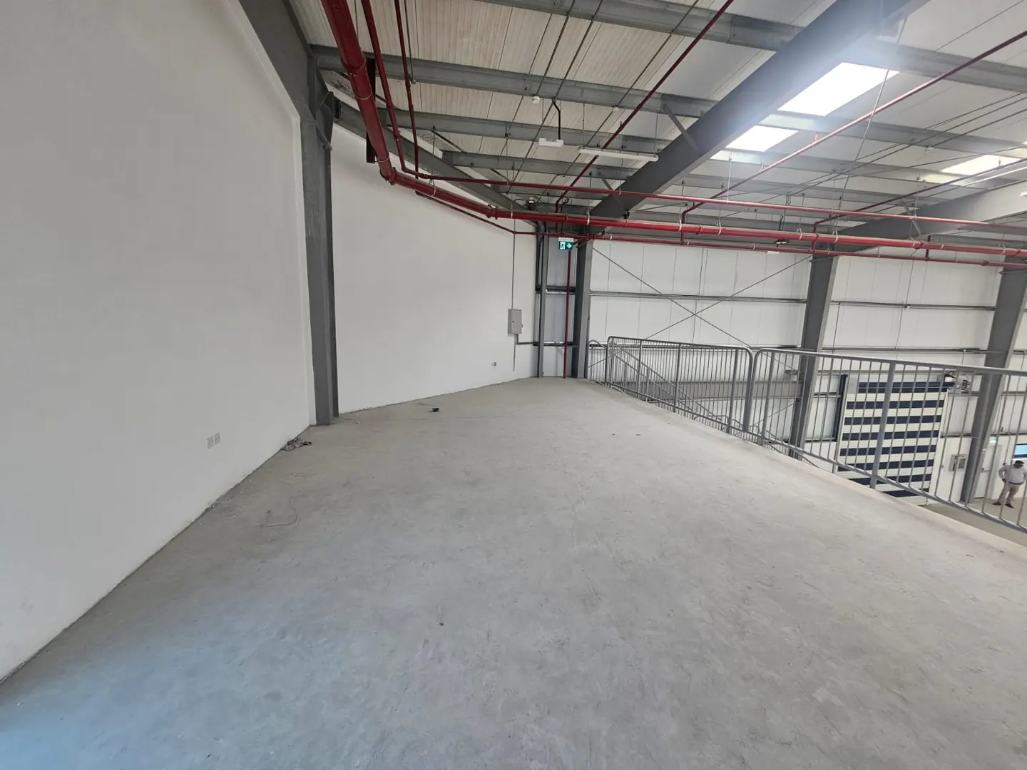 A large, empty warehouse space with white walls, a concrete floor, and a high ceiling with red pipes. A metal railing is visible on the right side.