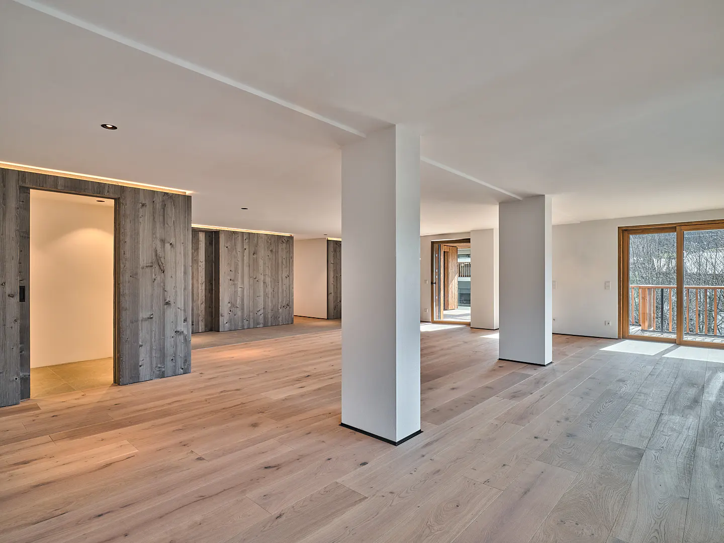 Spacious room with light wood floors, white walls, and gray wood accent walls. Large windows and white support columns.