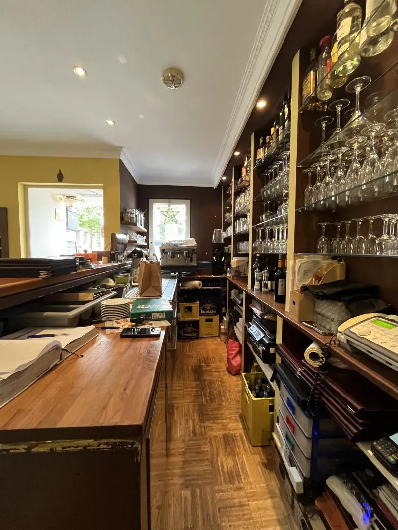 Interior view of a bar with a wooden counter, shelves stocked with bottles and glassware, and a herringbone-patterned floor.