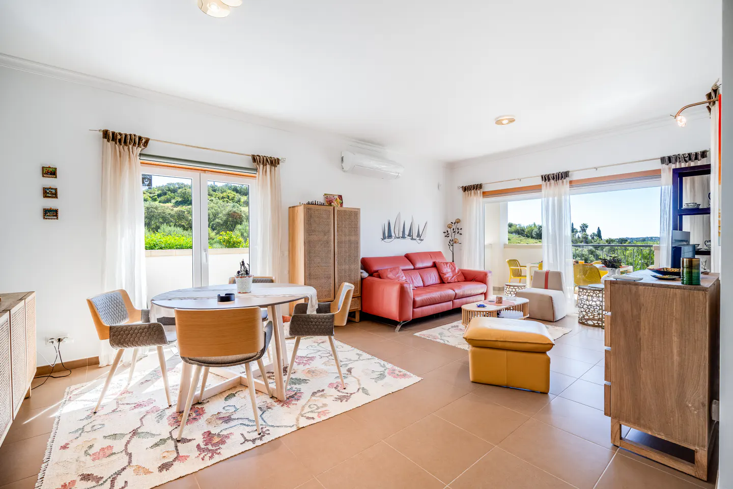 Bright living room with a red leather sofa, round table, and floral rug. Balcony doors open to a green landscape.