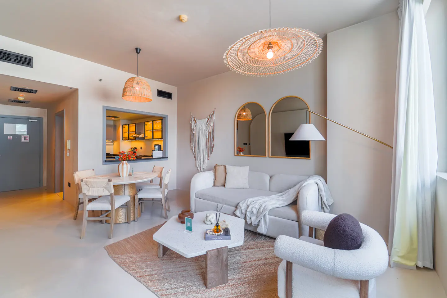Bright living room with white sofa, chair, and table on a jute rug. Rattan light fixtures hang above. Kitchen visible through opening.