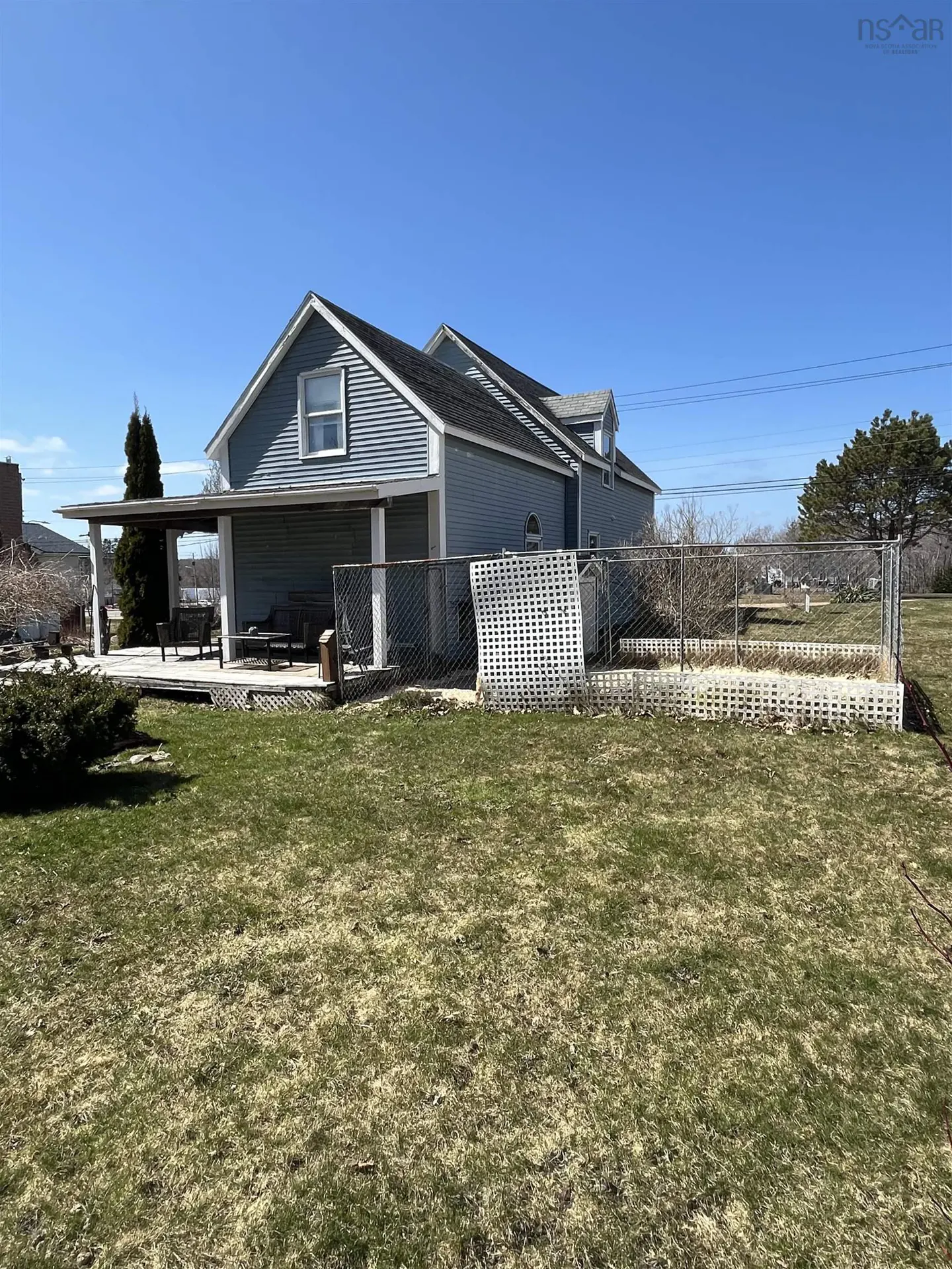 A light blue house with a covered porch and a black roof, surrounded by a green lawn and a chain-link fence.