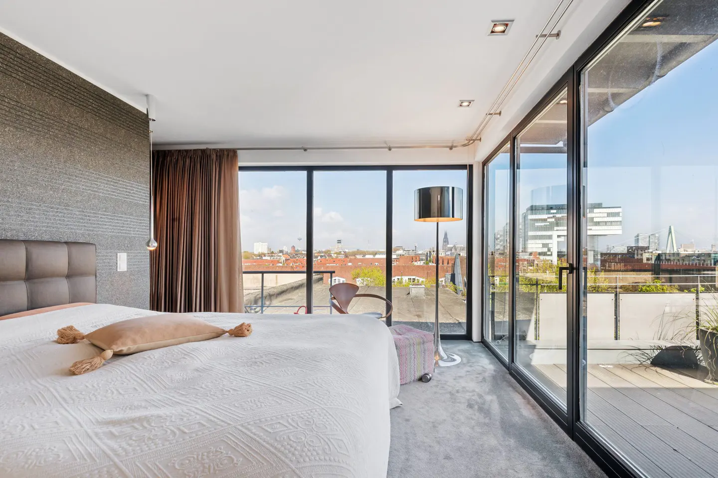 Bedroom with a white bed, brown headboard, and a large window with a city view. A silver floor lamp stands near the window.
