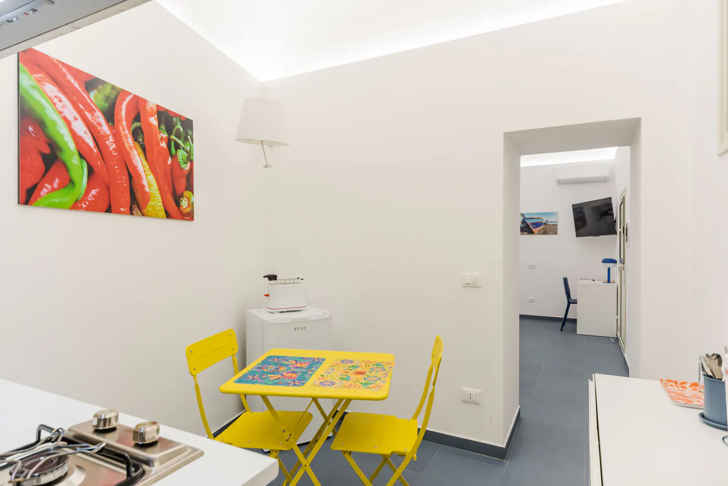 Bright kitchen with yellow table and chairs, white walls, and a colorful pepper print. A doorway leads to another room.