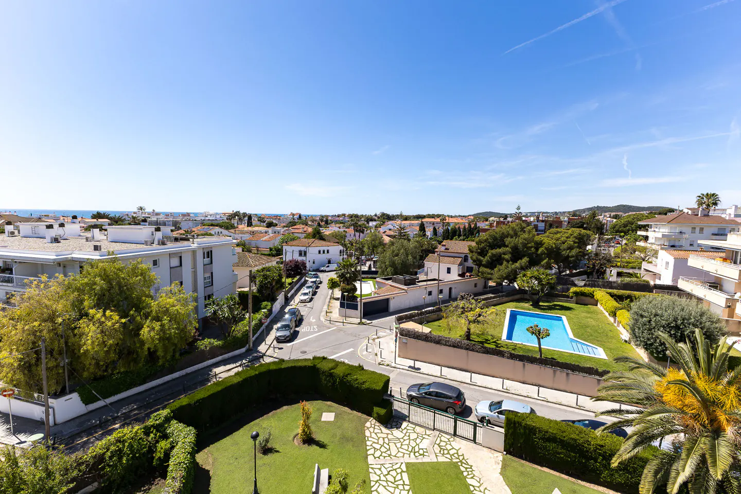 Aerial view of a residential neighborhood with white buildings, red tile roofs, green trees, and a blue swimming pool under a clear blue sky.
