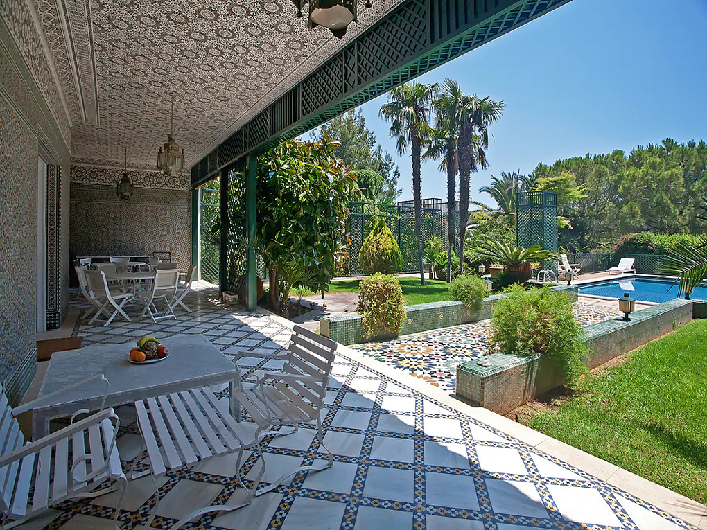 Covered patio with white furniture overlooks a pool and lush green garden with palm trees under a clear blue sky.