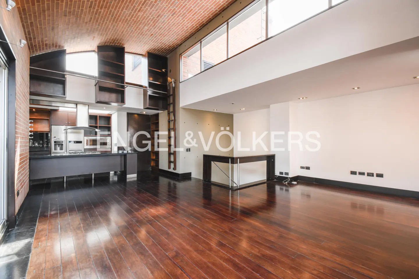 Open-concept loft with brick ceiling, hardwood floors, and a modern kitchen. Bookshelves and a ladder add character. Engel & Völkers logo visible.