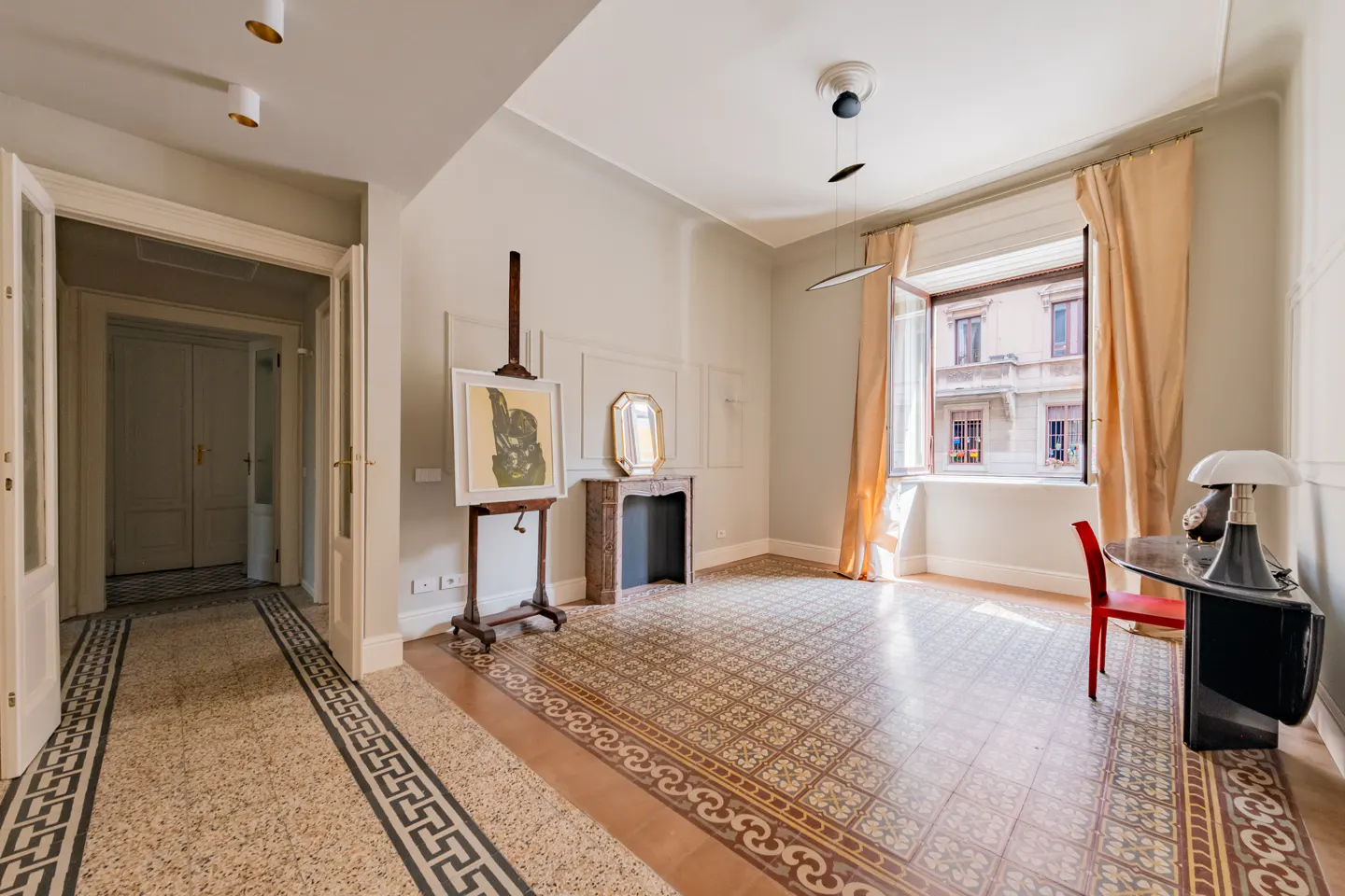 Bright room with patterned tile floor, easel with painting, fireplace, desk with lamp, and open window with peach curtains.