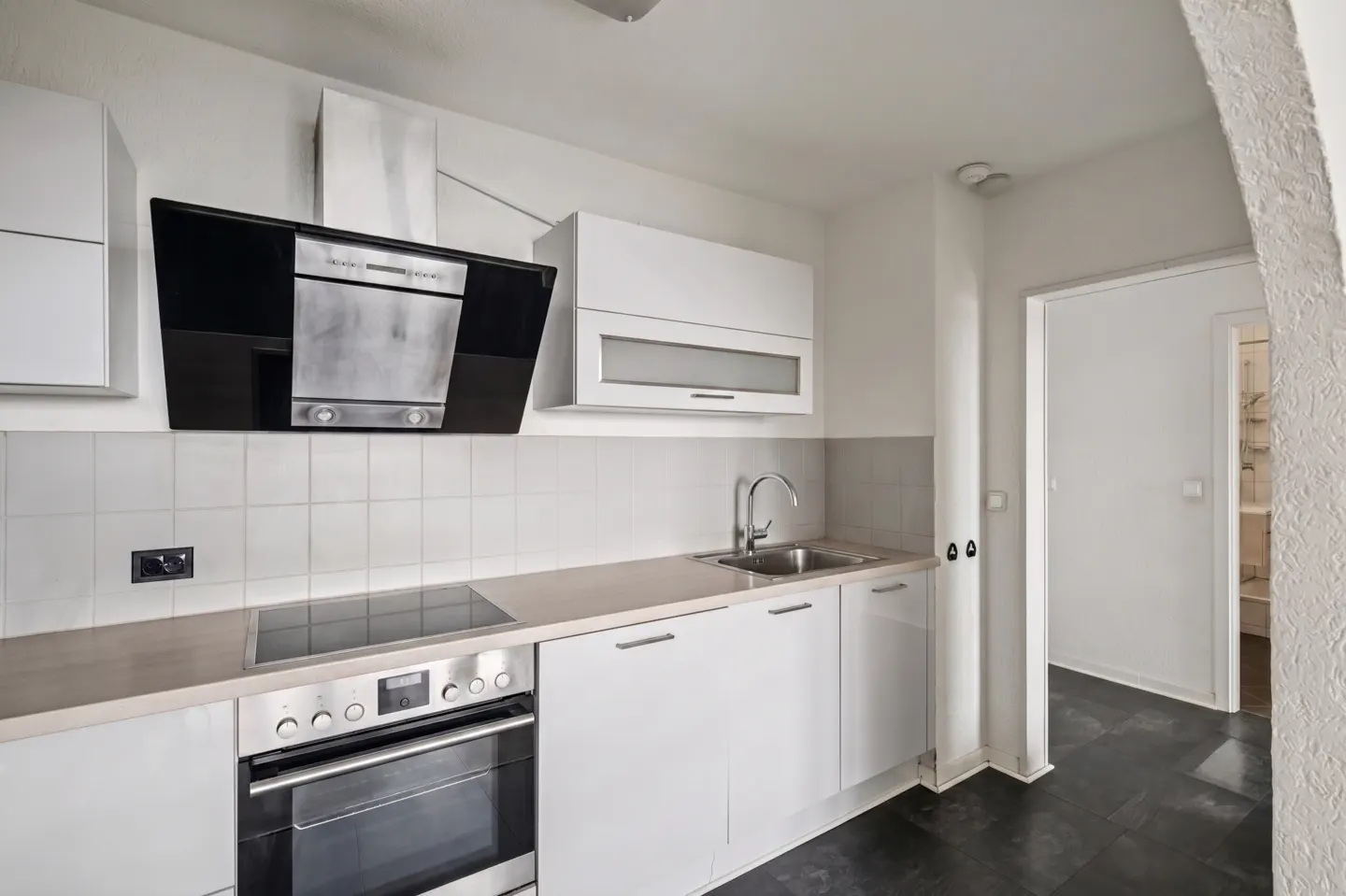 A modern kitchen with white cabinets, stainless steel appliances, and a black range hood. A doorway leads to another room.