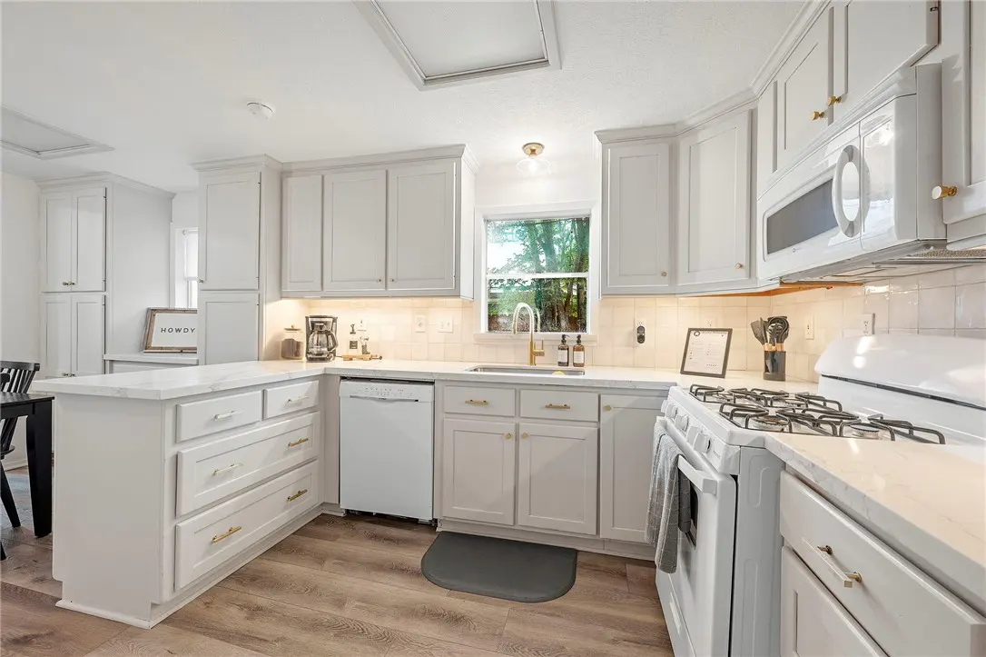 Bright kitchen with white cabinets, marble countertops, and wood floors. A window above the sink looks out to trees.