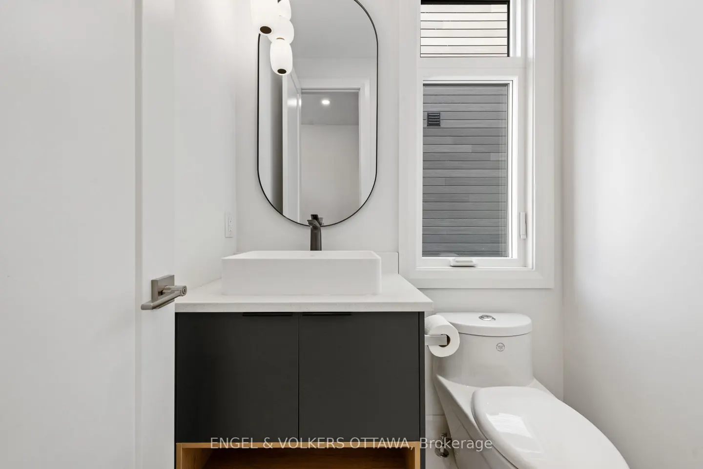 A modern powder room with white walls, a black vanity, a white sink, and an oval mirror. A toilet is next to the vanity.