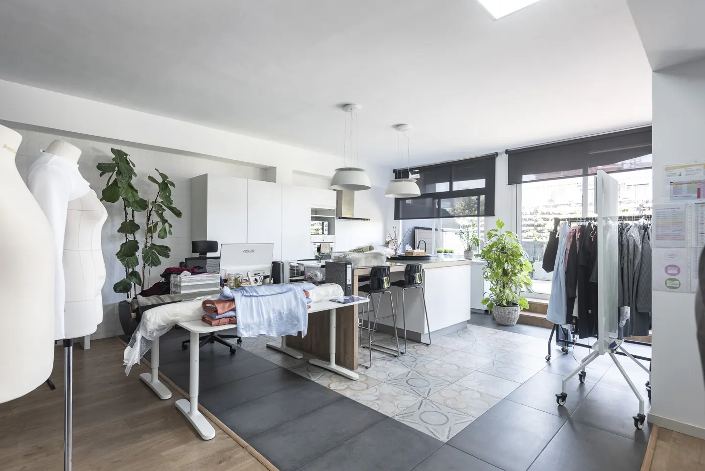 Bright, open studio with kitchen. Mannequins, plants, and a table with fabric are in the foreground. A clothing rack stands near a large window.