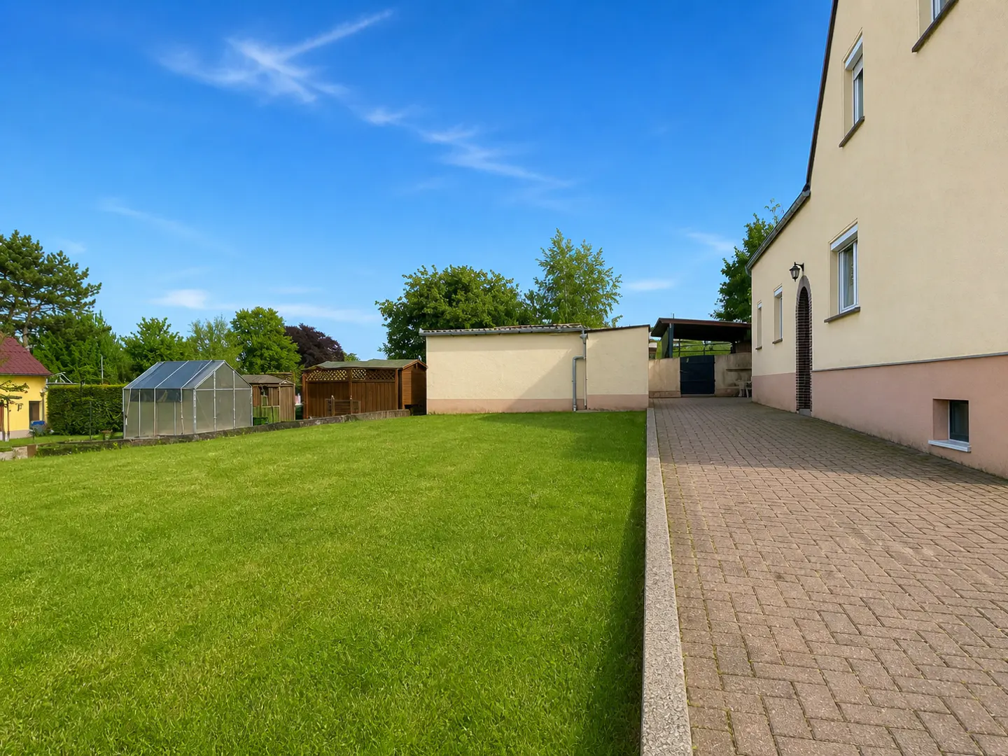A large green lawn next to a paved driveway leading to a light-colored house with a greenhouse in the background.