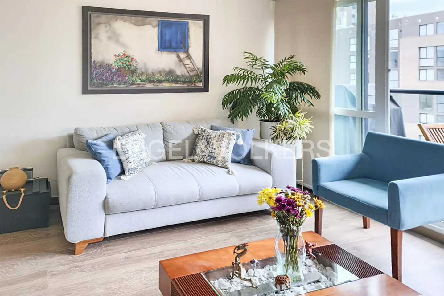 Living room with a gray sofa, blue chair, and a wooden coffee table with flowers. A painting hangs above the sofa. Balcony in the background.