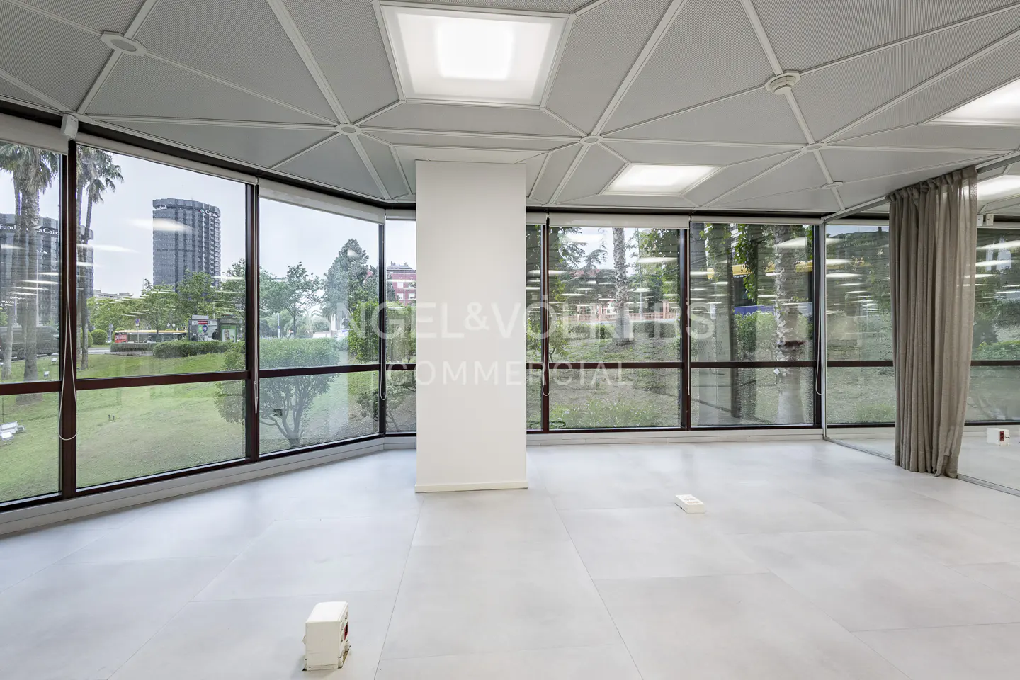 Bright, empty office space with large windows overlooking green space. Gray tile floor and geometric ceiling.