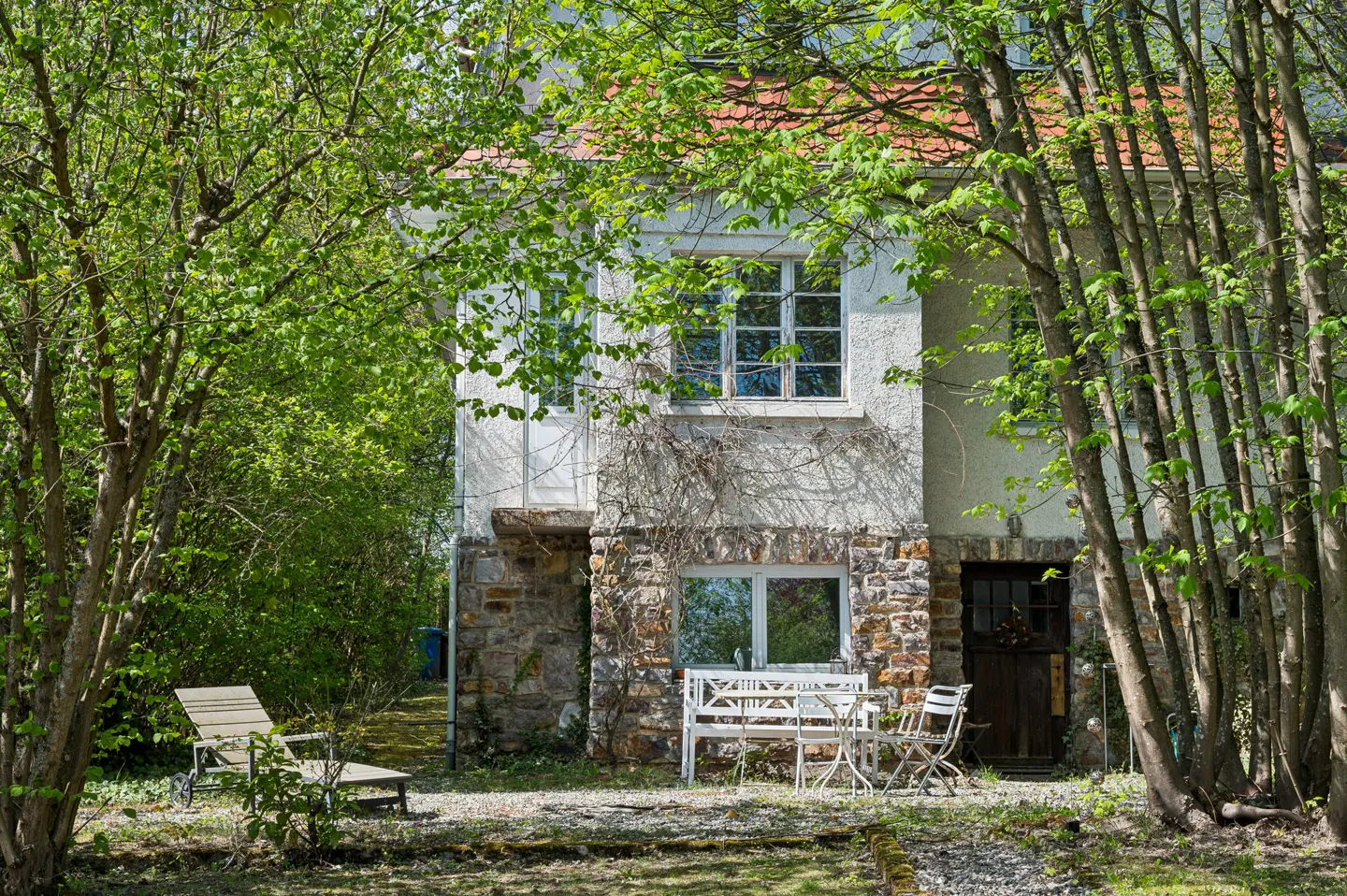 Two-story house with stone base, white stucco upper, red tile roof, surrounded by trees. White table and chairs sit outside.