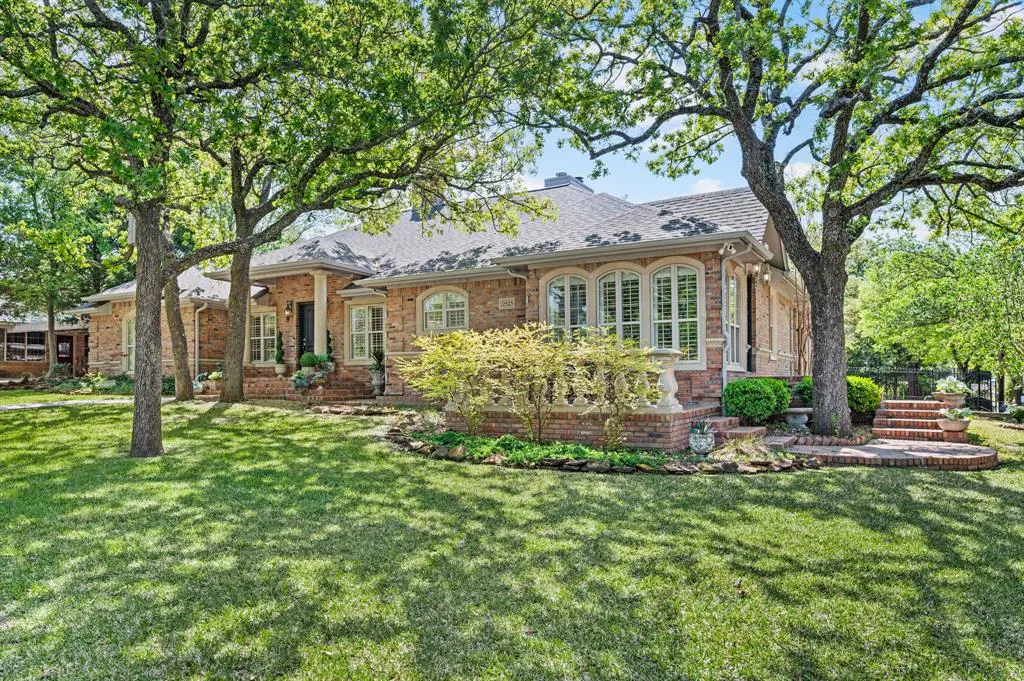 A single-story brick house with a gray roof is framed by large trees and a green lawn.