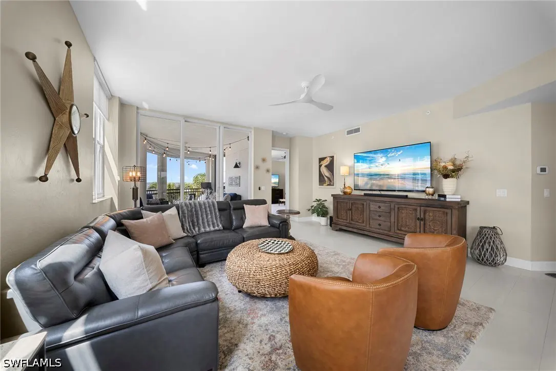 Bright living room with a gray leather sectional, brown leather chairs, and a woven coffee table on a patterned rug. A TV sits atop a carved wooden console.