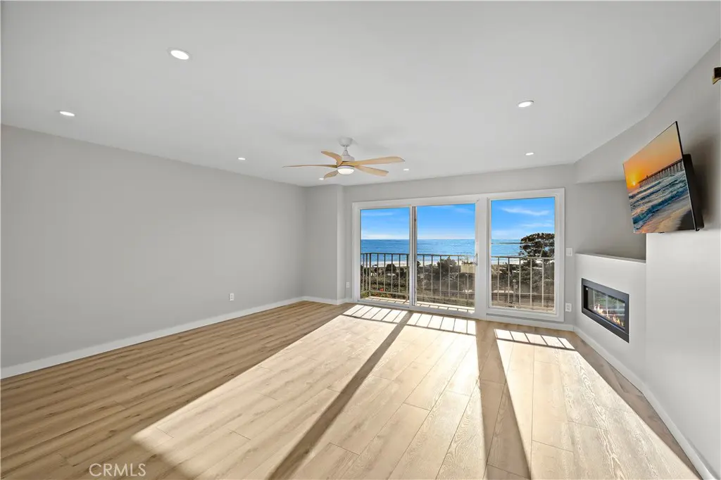 Bright living room with wood floors, gray walls, and ocean view through sliding glass doors. A ceiling fan and fireplace add comfort.