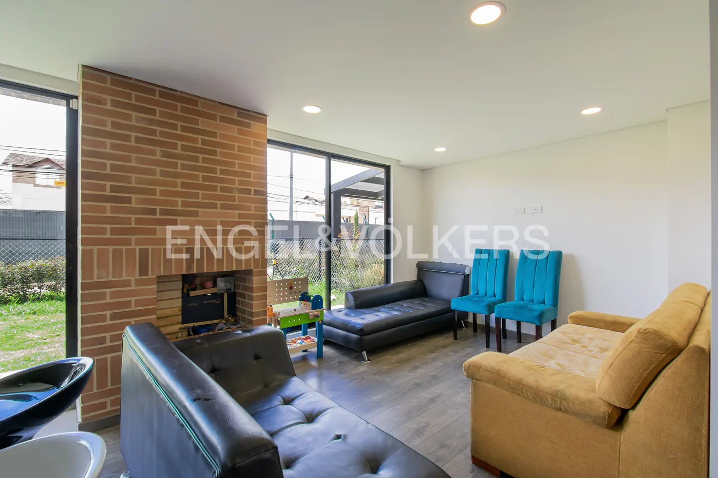 A bright living room with a brick fireplace, black leather couches, and two turquoise chairs. A yellow couch sits on a gray wood floor.
