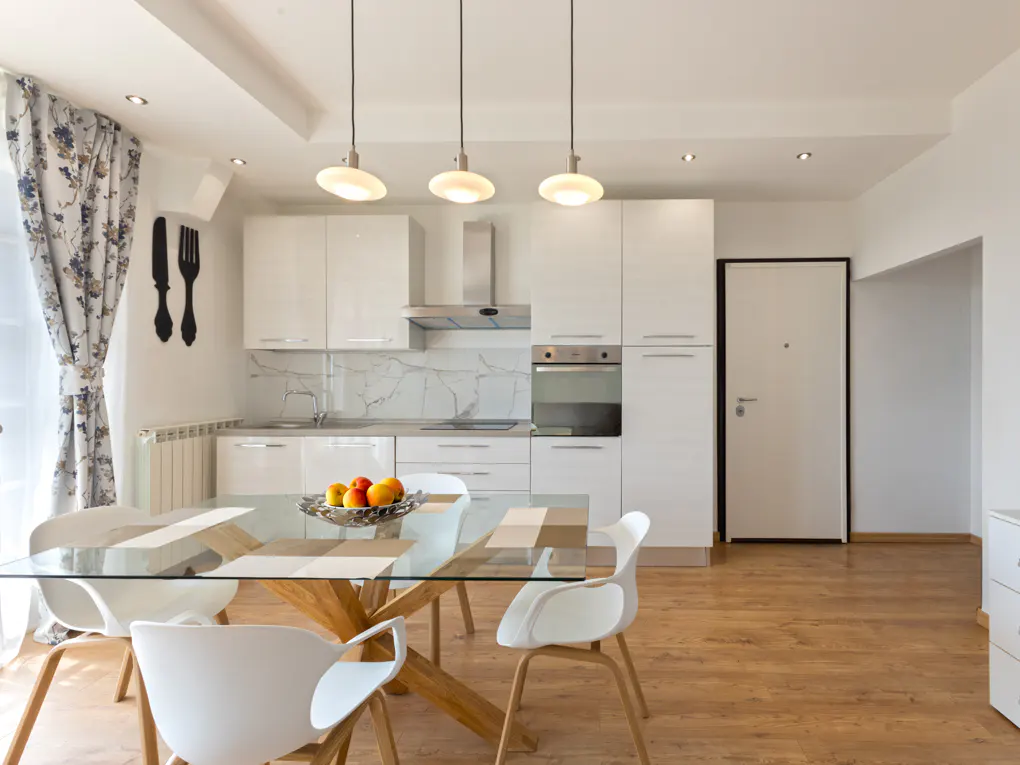 Bright, modern kitchen with white cabinets, wood floors, and a glass table with fruit. Floral curtains and utensil art add charm.