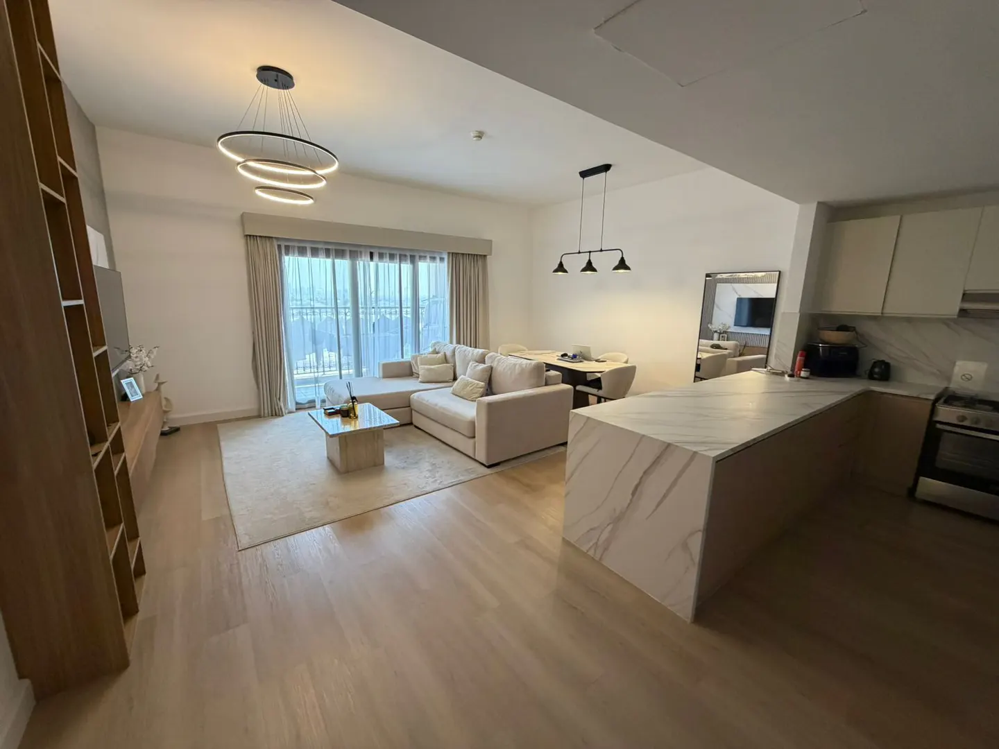 A bright, modern apartment interior with a beige sectional sofa, marble kitchen island, and wood floors. A dining area is visible in the background.