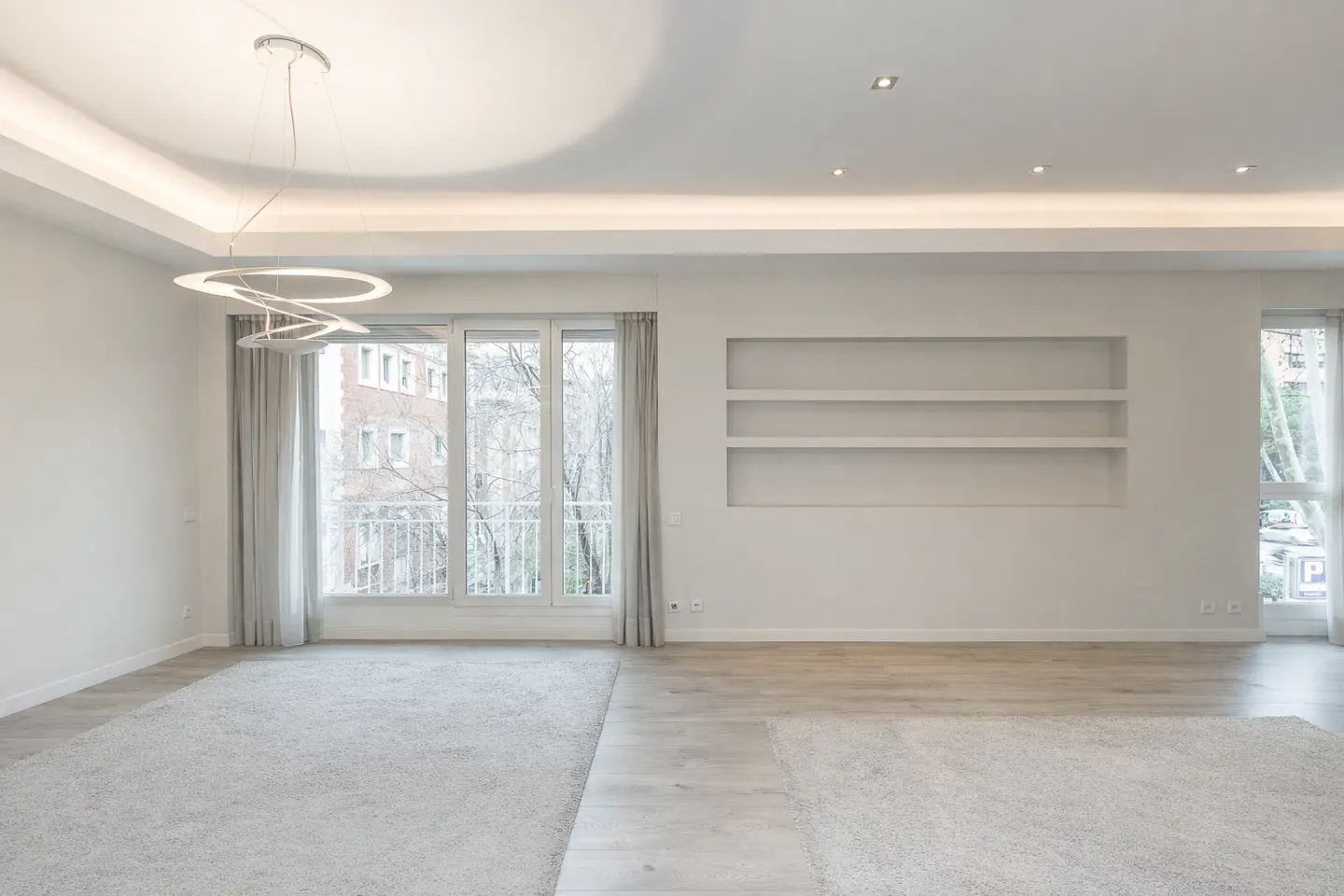 Bright, empty living room with white walls, light wood floors, and a gray rug. Large windows offer a view of a building and trees. A modern chandelier hangs from the ceiling.