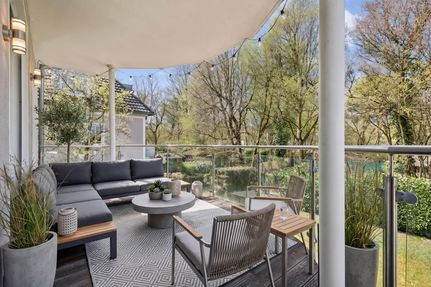 A modern outdoor patio with gray sectional sofa, round concrete table, and chairs on a patterned rug, overlooking lush green trees.