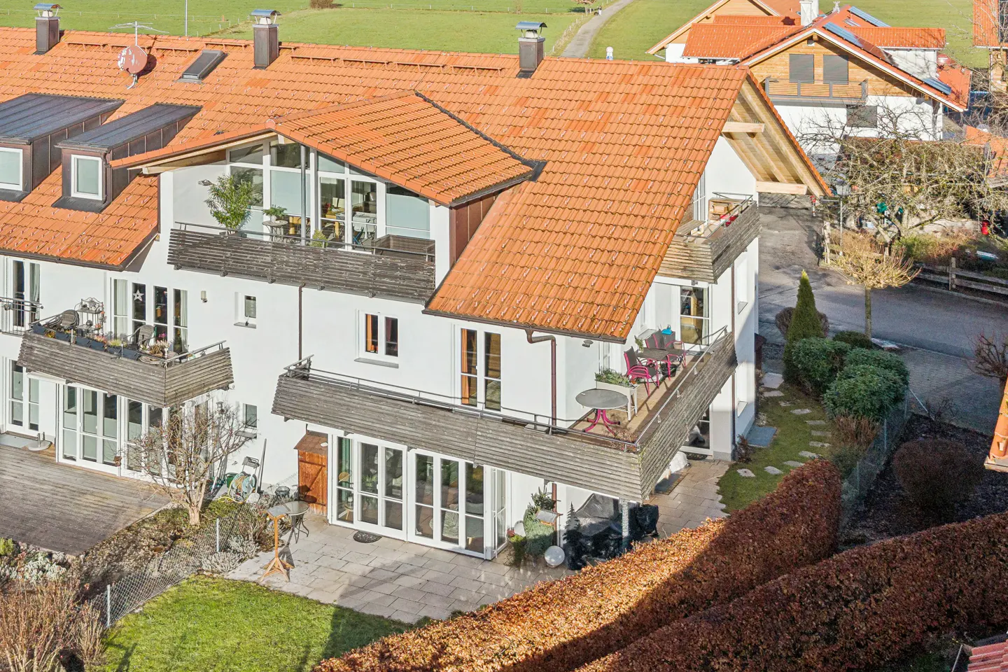 Aerial view of a white, multi-story house with orange tile roof and balconies, surrounded by green grass and trees.