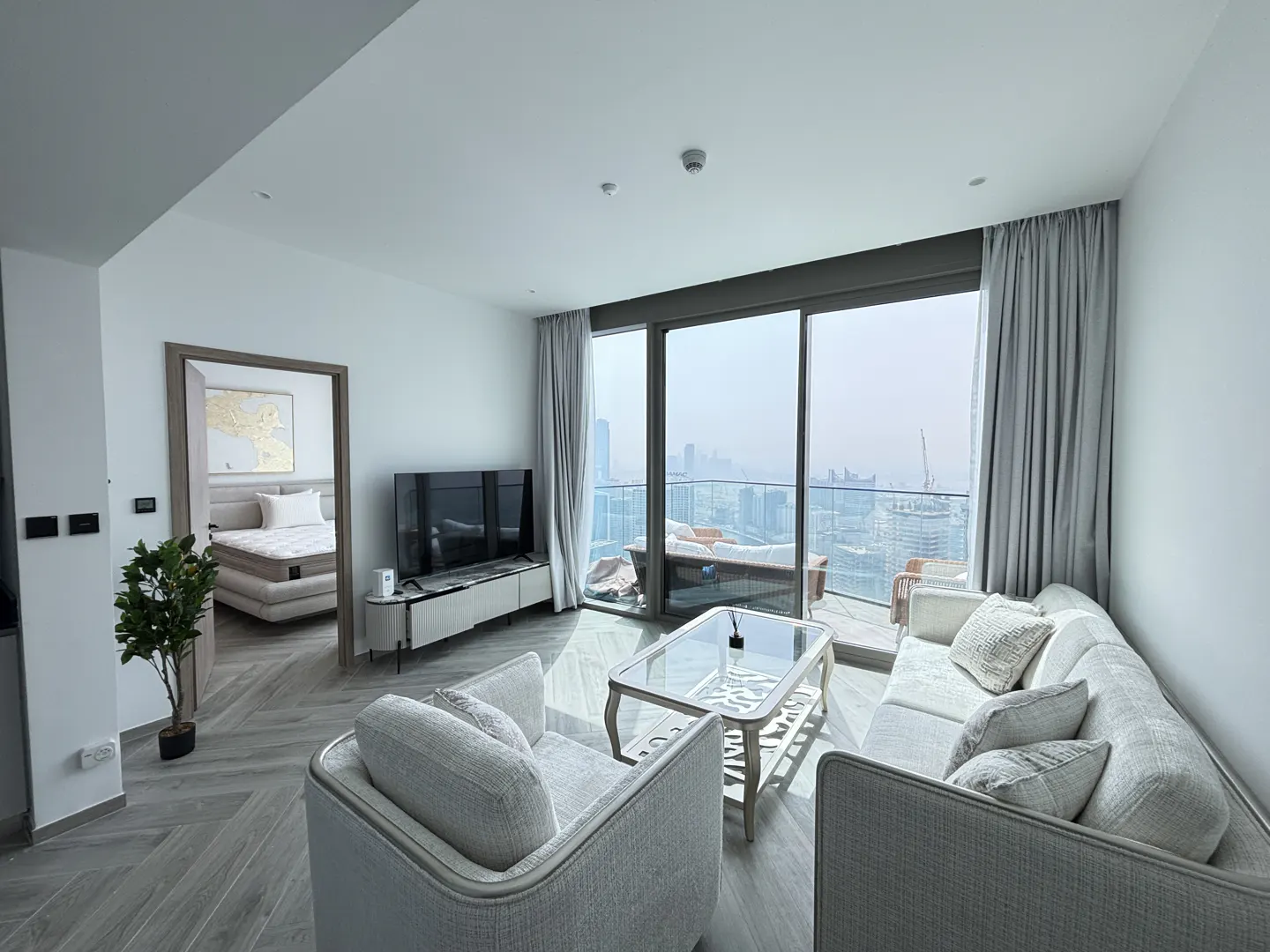 Bright, modern living room with gray sofa, chairs, and glass table. Balcony with city view. Bedroom visible through doorway.
