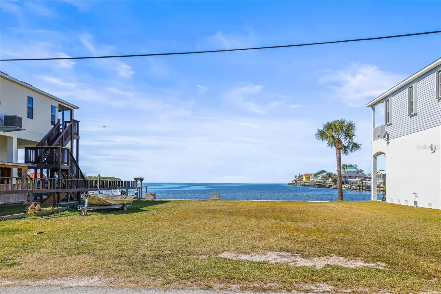Waterfront property with a grassy lot between two houses, leading to a blue bay under a partly cloudy sky. A palm tree stands on the right.