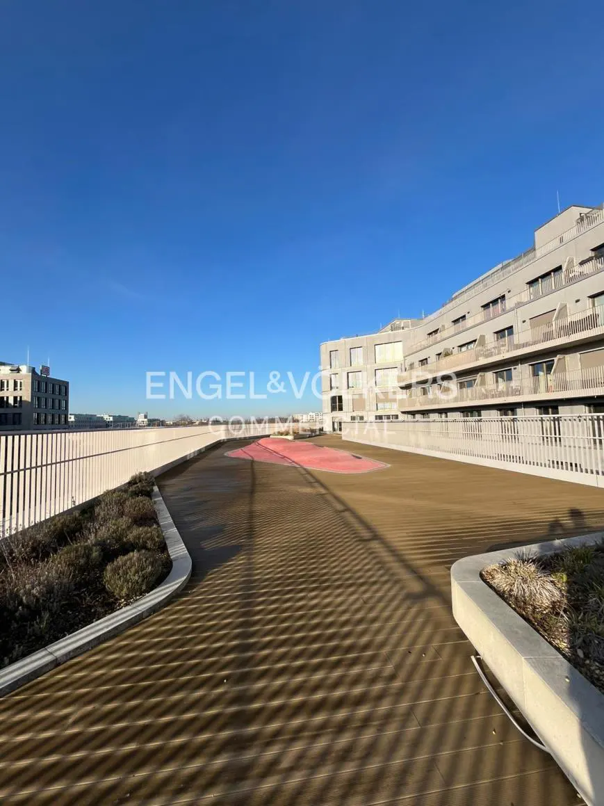 Rooftop view of a modern building with a wooden deck, white railings, and greenery. Clear blue sky above. Engel & Volkers logo visible.