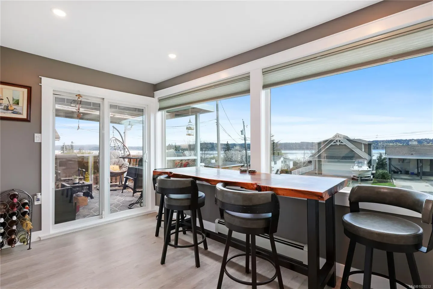 Bright kitchen with a long wooden countertop, gray stools, and a large window overlooking a waterfront view. Sliding glass doors lead to a patio.