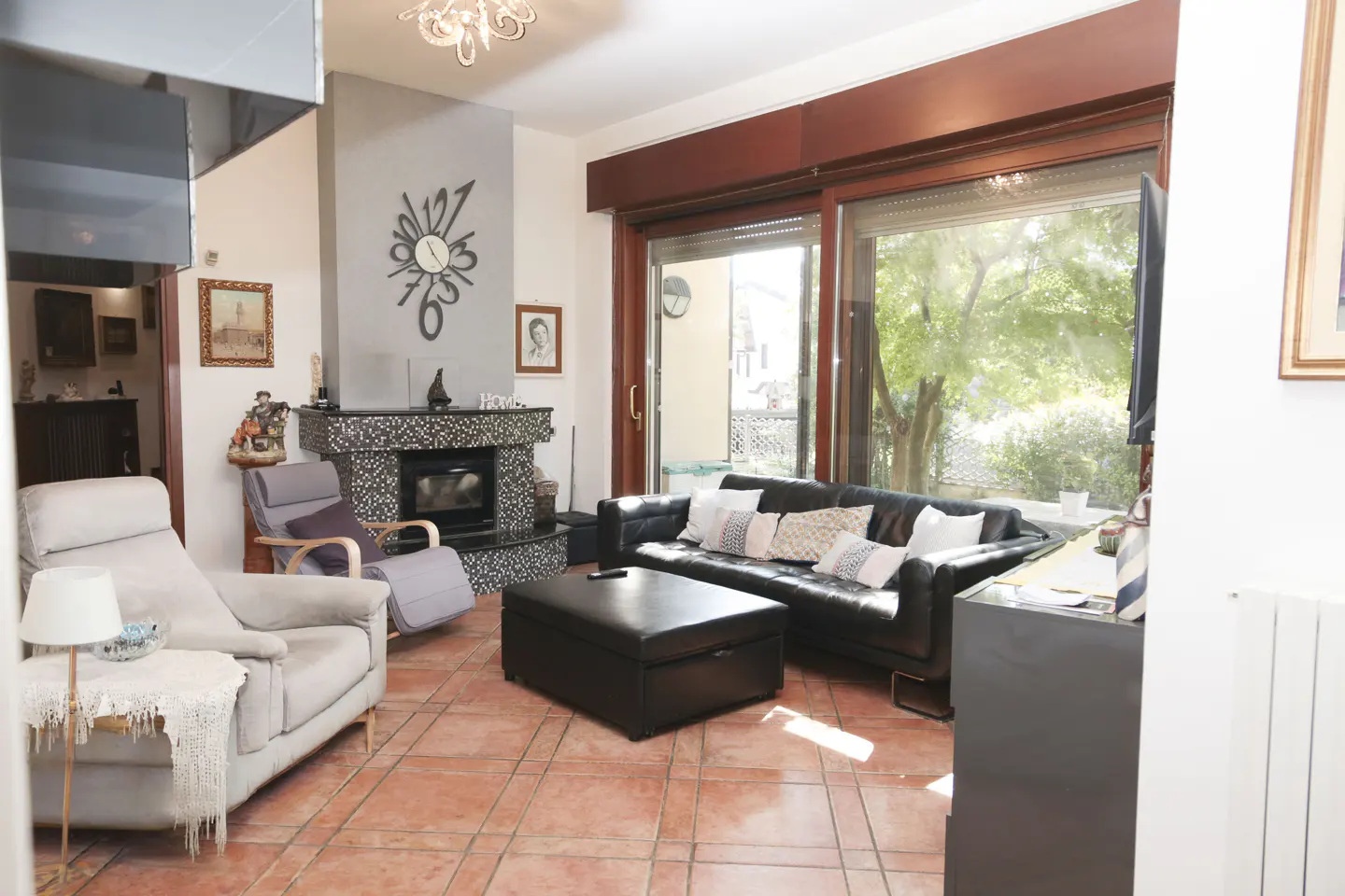A living room with terracotta tile flooring, a black leather sofa, and a mosaic fireplace. Large windows overlook a green outdoor space.