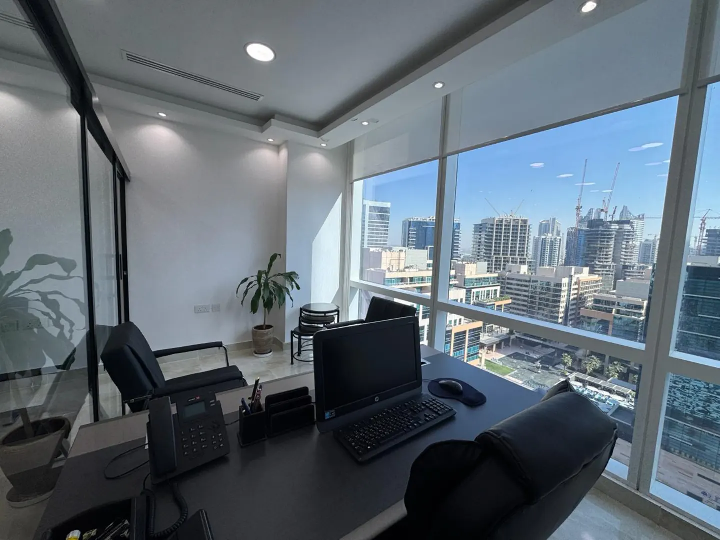 Modern office with a desk, computer, and black chairs, featuring floor-to-ceiling windows with a city skyline view.