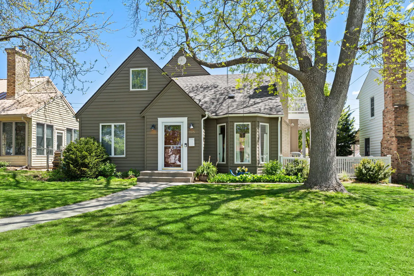 A one-and-a-half story olive green house with a gray roof and a large tree in the front yard.