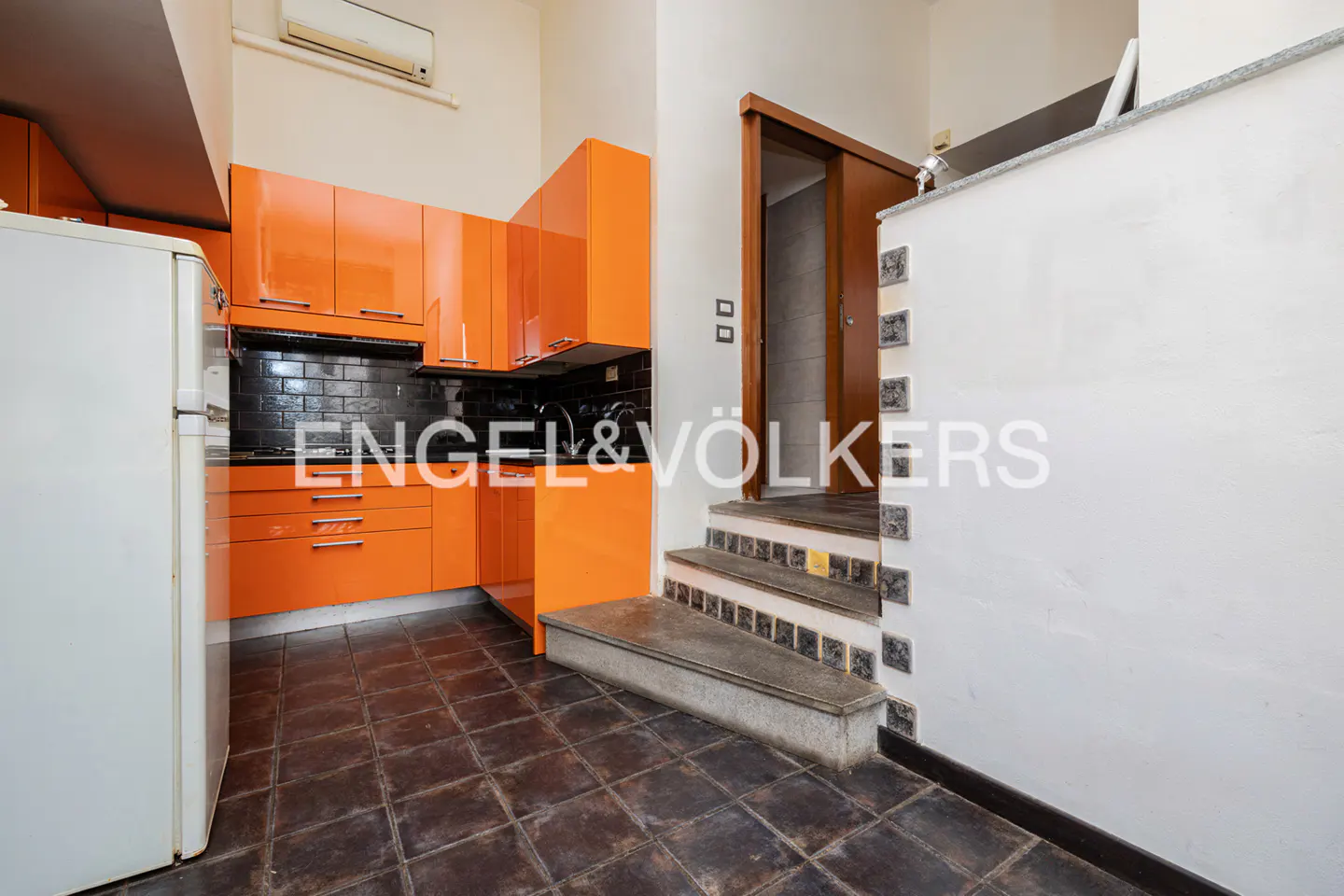 A kitchen with orange cabinets, black backsplash, and a white refrigerator. Stairs with decorative tiles lead to a doorway. Dark tiled floor.