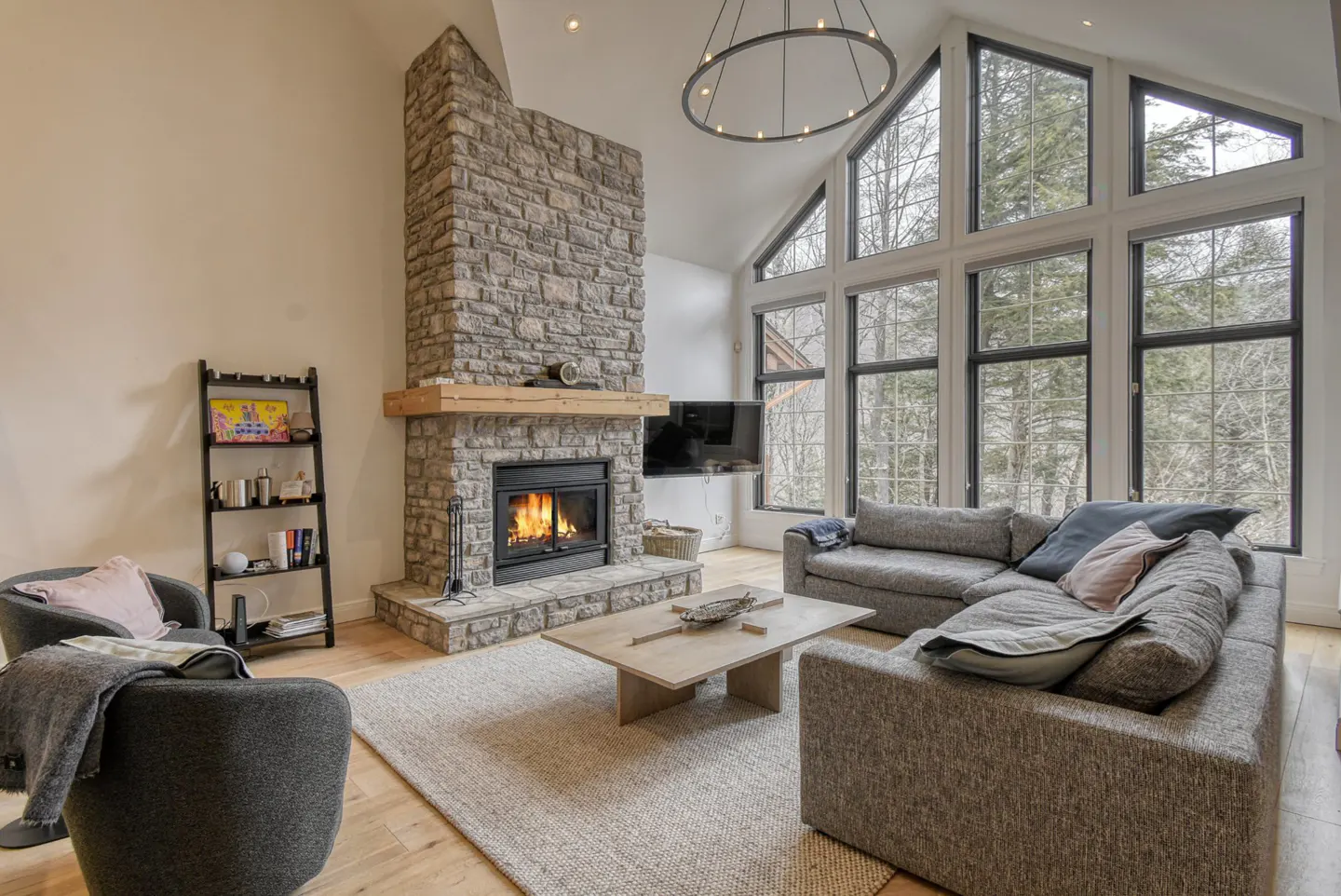Living room with stone fireplace, gray sofa, and large windows overlooking trees. A round chandelier hangs from the ceiling.