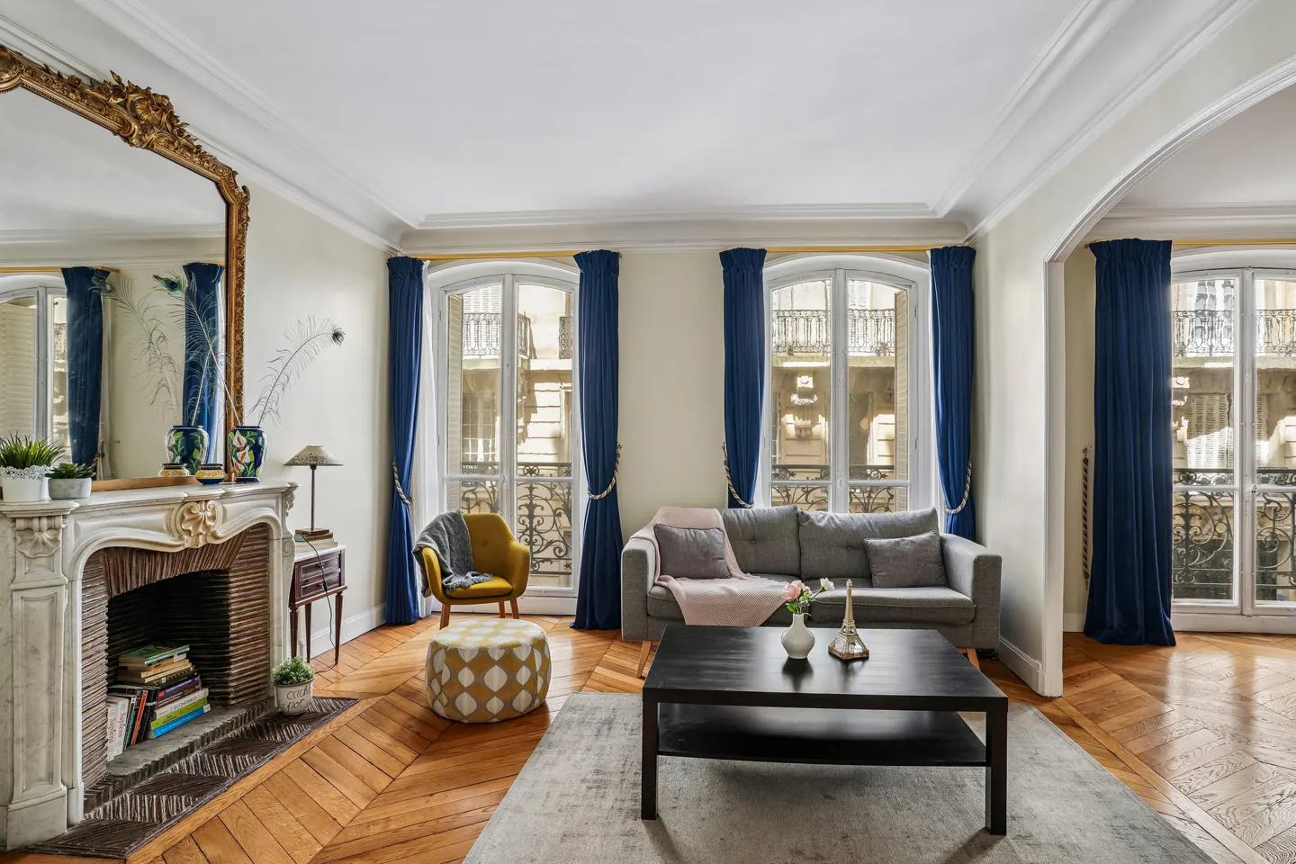 Living room with hardwood floors, fireplace, and large windows with blue curtains. A gray sofa and black coffee table sit on a gray rug.
