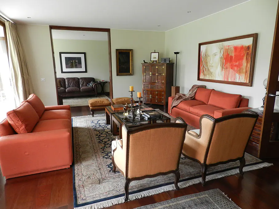 Living room with orange sofas, armchairs, and a dark wood floor. A doorway leads to another room with a brown leather sofa.